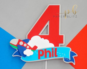 May include: Red and white number four cake topper with a blue and red airplane flying through a white cloud. The topper also has a blue banner with the name "Phil" written in red.