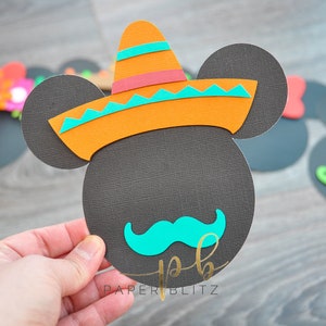 以下が含まれることがあります： 黒色の紙で作ったミッキーマウスのクラフトで、カラフルなソンブレロとターコイズブルーの口ひげを付けています。クラフトは手で持たれており、下部に「PAPERBLITZ」というテキストが表示されています。