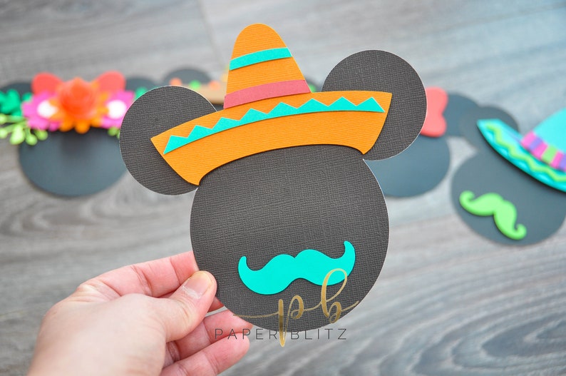 以下が含まれることがあります： 黒色の紙で作ったミッキーマウスのクラフトで、カラフルなソンブレロとターコイズブルーの口ひげを付けています。クラフトは手で持たれており、下部に「PAPERBLITZ」というテキストが表示されています。