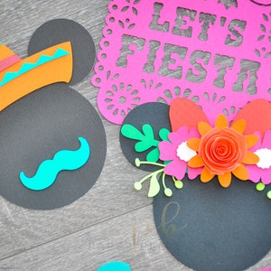 以下が含まれることがあります： 黒色の紙でできたミッキーマウスの耳が2つ。片方の耳にはソンブレロと口ひげ、もう片方の耳には花飾り。背景は木目調の表面。上の看板には「Let's Fiesta」と書かれている。