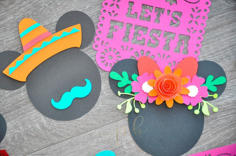 以下が含まれることがあります： 黒色の紙でできたミッキーマウスの耳が2つ。片方の耳にはソンブレロと口ひげ、もう片方の耳には花飾り。背景は木目調の表面。上の看板には「Let's Fiesta」と書かれている。