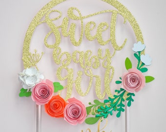 May include: A gold glitter "Sweet Baby Girl" cake topper with a floral design. The topper features a circular frame with the text in elegant script. It is adorned with paper flowers in shades of pink, orange, and white, with green leaves and blue accents.