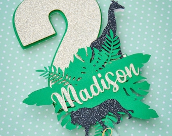 May include: A jungle-themed birthday cake topper featuring a glittery gold number 2 with green trim. The topper includes a giraffe, lion, and the name "Madison" in a cursive font, all set against green tropical leaves.
