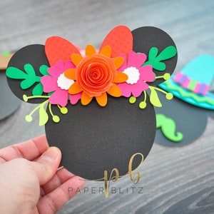 以下が含まれることがあります： オレンジ、ピンク、白、緑の紙の花で作られた花輪が付いた黒い紙のミッキーマウスの耳。花輪には赤いリボンも付いています。耳には金色の文字で「PAPER BLITZ」と書かれています。