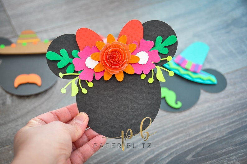 以下が含まれることがあります： オレンジ、ピンク、白、緑の紙の花で作られた花輪が付いた黒い紙のミッキーマウスの耳。花輪には赤いリボンも付いています。耳には金色の文字で「PAPER BLITZ」と書かれています。