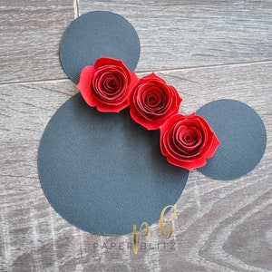 以下が含まれることがあります： 黒い紙のミッキーマウスの頭のシルエットに、赤い紙のバラが3つ並んでいます。シルエットは木目調の背景の上にあります。画像の下部に「PAPER BLITZ」というテキストが表示されています。