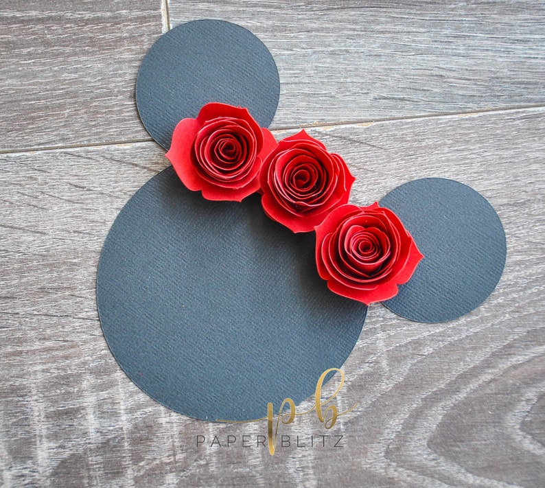 以下が含まれることがあります： 黒い紙のミッキーマウスの頭のシルエットに、赤い紙のバラが3つ並んでいます。シルエットは木目調の背景の上にあります。画像の下部に「PAPER BLITZ」というテキストが表示されています。