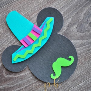 以下が含まれることがあります： 木目調の背景に、グレーの紙でできたミッキーマウスの頭の形をしたクラフト。青い帽子と緑のひげが付けられている。