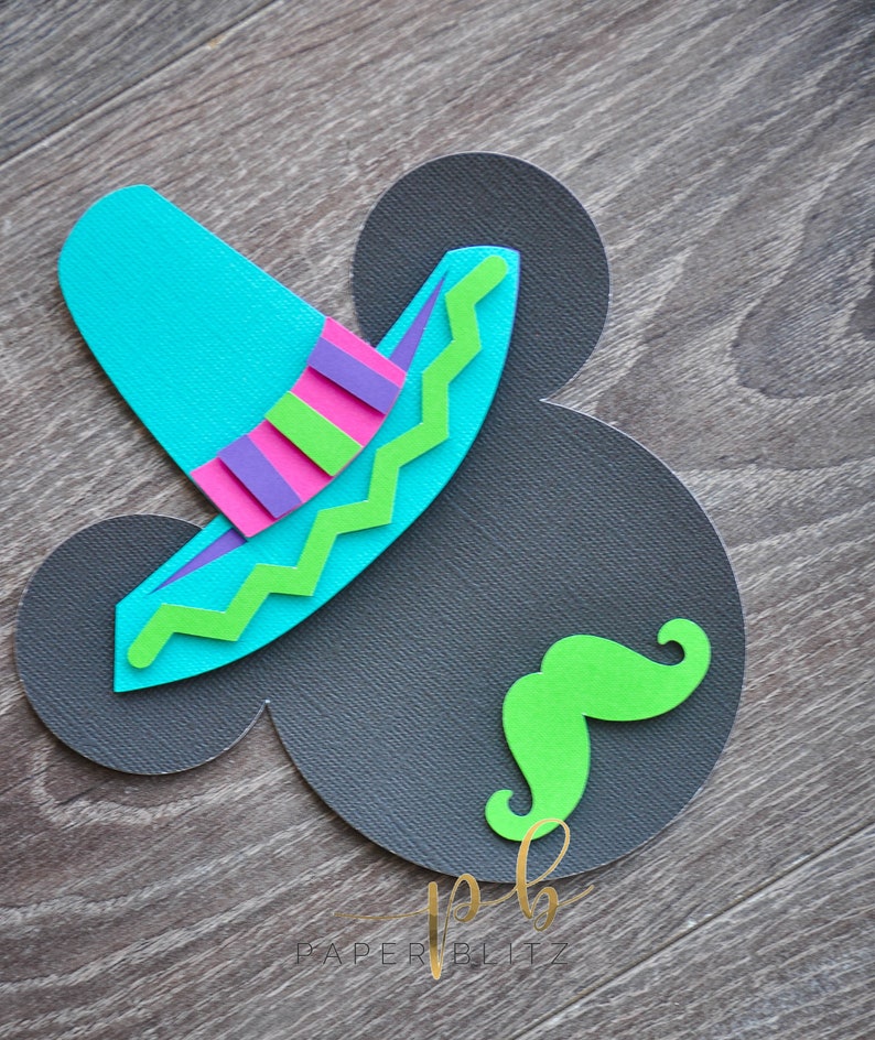 以下が含まれることがあります： 木目調の背景に、グレーの紙でできたミッキーマウスの頭の形をしたクラフト。青い帽子と緑のひげが付けられている。