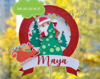May include: Red and green Christmas cake topper with a Santa Claus figure holding a Christmas tree. The topper has a white banner with the name "Maya" and the text "Shake and light me up!" in a speech bubble.
