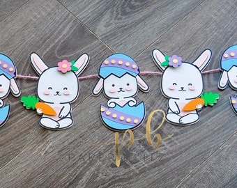 May include: A decorative Easter banner featuring cartoon bunnies and Easter eggs. The bunnies are white with pink accents, holding carrots, and emerging from blue and purple eggs. The banner is strung on a pink string, perfect for Easter decorations.