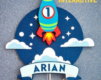 May include: A blue, red, and green rocket ship cake topper with the number 1, clouds, stars, and the name "ARIAN" on a banner. The word "INTERACTIVE" is printed in yellow. The background is a glittery blue.