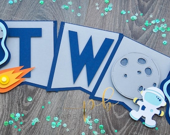 May include: A blue and gray paper banner with the word "TWO" in blue letters. The banner is decorated with rockets, stars, and moons. It is perfect for a space-themed birthday party.