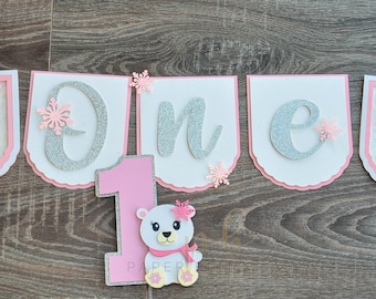 May include: A decorative banner for a first birthday celebration. The banner spells "ONE" in silver glitter letters on white pennants, accented with pink snowflakes. Two pennants feature a polar bear with a pink scarf and flower. A pink number "1" and polar bear are also included.