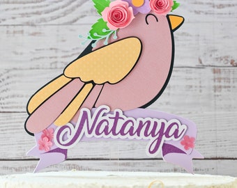 May include: A pink paper bird cake topper with a yellow wing, a purple banner with the name "Natanya" and a floral crown with pink and purple flowers.