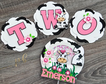May include: A set of birthday decorations featuring a cow theme. Includes the word "TWO" in pink letters, a cow-themed shaker, and a personalized name. The decorations are adorned with pink flowers and cow print accents.