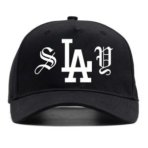 Puede incluir: Gorra de béisbol negra con el texto blanco "S LA Y" en una fuente estilizada. La gorra tiene una visera curva y un botón en la parte superior. Un accesorio casual clásico.
