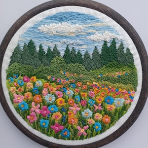 Könnte beinhalten: Handgestickte Stickerei in einem runden Holzrahmen. Die Szene zeigt eine lebendige Wiese mit bunten Blumen, grünem Gras und immergrünen Bäumen unter blauem Himmel mit weißen Wolken. Ein dekoratives Stück für die Wanddekoration.