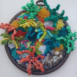 May include: A circular embroidery hoop art piece featuring a vibrant, textured coral reef scene. The design incorporates various shades of green, orange, yellow, and blue yarn, with raised stitches, pom-poms, and small beads to mimic underwater life.