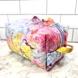 May include: A quilted, rectangular cosmetic bag with a vibrant, colorful batik pattern. The bag features a lavender zipper, a small yellow handle, and a bow-shaped accent. The fabric showcases a mix of pink, yellow, and purple hues.