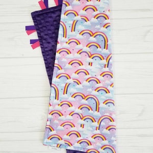 Op de afbeelding: Een paarse en witte babydeken met een regenboog- en hartpatroon. De deken heeft aan de ene kant een zachte, minky textuur en aan de andere kant een gladde, katoenen textuur. Het heeft paarse satijnen lintjes.
