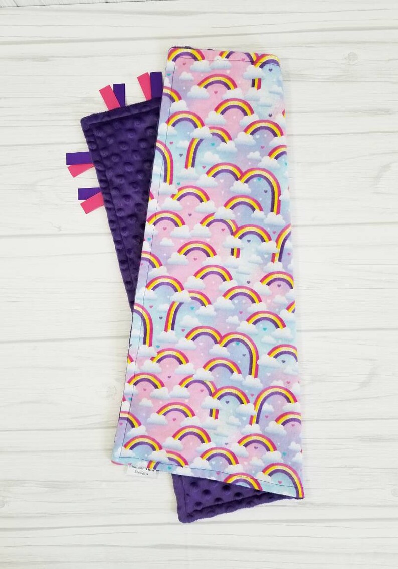 Op de afbeelding: Een paarse en witte babydeken met een regenboog- en hartpatroon. De deken heeft aan de ene kant een zachte, minky textuur en aan de andere kant een gladde, katoenen textuur. Het heeft paarse satijnen lintjes.