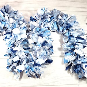 May include: Three fabric garlands in shades of blue and white, arranged on a light wood surface. The garlands are made of strips of fabric tied together, creating a textured, decorative element. The fabric has various patterns.