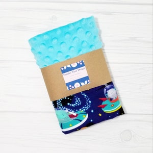 May include: A blue and white minky baby blanket with a space theme. The blanket features a dark blue background with white stars, and a variety of colorful spaceships and aliens. The blanket is wrapped in brown paper with a label that reads "Shudder Time Designs".