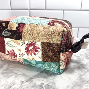 May include: A quilted fabric cosmetic bag with a patchwork design. The bag features a variety of floral and geometric patterns in shades of burgundy, teal, beige, and brown. It has a zipper closure and a fabric pull.
