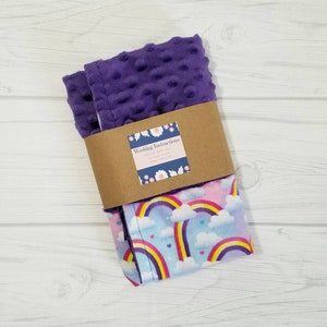 Op de afbeelding: Een paarse en roze babydeken met een regenboog- en hartprint. De deken is dubbelgevouwen en heeft een bruin label met de tekst "Washing Instructions".