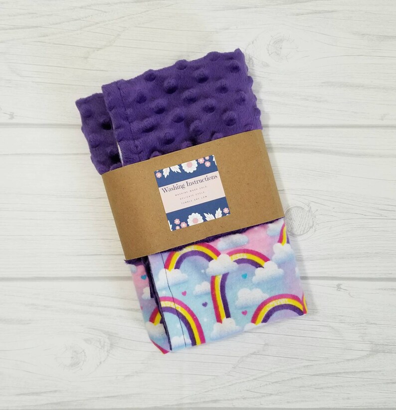 Op de afbeelding: Een paarse en roze babydeken met een regenboog- en hartprint. De deken is dubbelgevouwen en heeft een bruin label met de tekst "Washing Instructions".