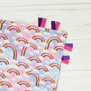 Op de afbeelding: Een zacht, roze en blauw babydekentje met een regenboog- en wolkenpatroon. Het dekentje heeft paarse en roze satijnen lintjes.