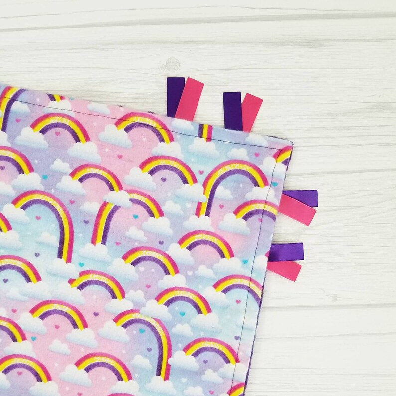 Op de afbeelding: Een zacht, roze en blauw babydekentje met een regenboog- en wolkenpatroon. Het dekentje heeft paarse en roze satijnen lintjes.