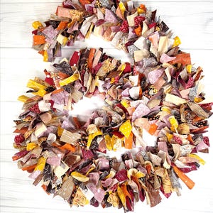 May include: A handmade wreath composed of fabric strips in various colors, including orange, yellow, red, and brown. The wreath is arranged in a circular shape, with the fabric strips creating a textured, layered effect. The background is a white wooden surface.
