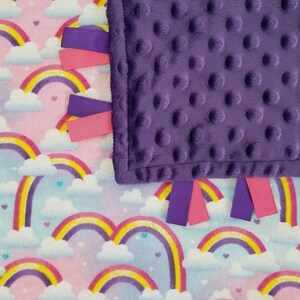 Op de afbeelding: Een paarse minky babydeken met regenboog- en wolkprint aan de ene kant en een zachte, gestructureerde paarse minky stof aan de andere kant. De deken heeft roze en paarse satijnen lintjes.