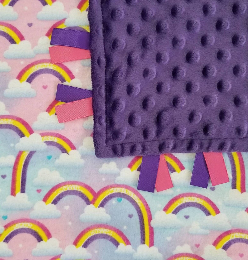 Op de afbeelding: Een paarse minky babydeken met regenboog- en wolkprint aan de ene kant en een zachte, gestructureerde paarse minky stof aan de andere kant. De deken heeft roze en paarse satijnen lintjes.