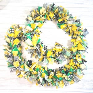 May include: A handmade wreath with a vibrant mix of fabrics in yellow, green, gray, and black. The wreath features a variety of patterns, including polka dots, gingham, and floral designs. The wreath is perfect for adding a touch of color and texture to any space.