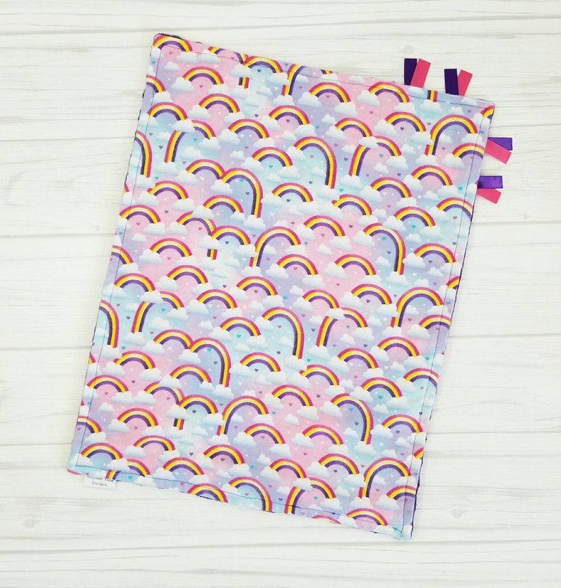 Op de afbeelding: Een zacht, roze en blauw babydekentje met een regenboog- en hartjespatroon. Het dekentje heeft paarse, roze en blauwe lintjes.