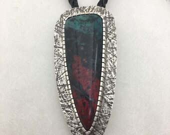 May include: A bolo tie featuring a large, shield-shaped pendant with a polished stone. The stone displays a mix of deep red, teal, and black hues, set in a textured silver frame. The tie has a braided black leather cord.