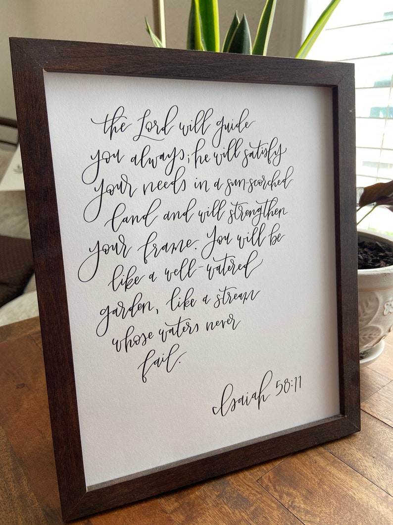 May include: A framed print with a black and white calligraphy quote from the Bible, Isaiah 58:11. The quote reads: "The Lord will guide you always; he will satisfy your needs in a sun-scorched land and will strengthen your frame. You will be like a well-watered garden, like a stream whose waters never fail."