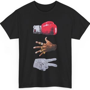 Peut inclure: T-shirt noir avec un motif graphique repr&eacute;sentant un gant de boxe rouge, une main orn&eacute;e de bagues dor&eacute;es et une main faisant le signe de la paix. Un design unique.