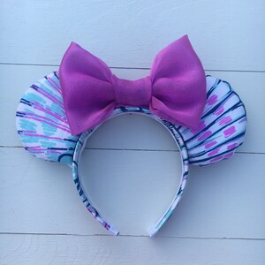May include: A headband with mouse ears and a large bow. The ears feature a white background with blue and purple abstract patterns. The bow is a solid, vibrant purple. The headband is white with a matching pattern.