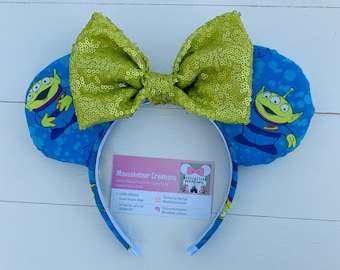 May include: Blue mouse ears featuring the Little Green Men from Toy Story. The ears have a blue background with a pattern of white dots and the aliens. A large, sequined, lime green bow sits between the ears. A business card is visible.