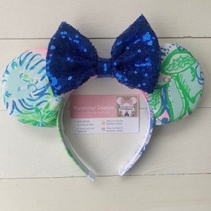 May include: A pair of mouse ears with a blue sequin bow and patterned ear covers. The ear covers feature a colorful floral design in shades of blue, green, and pink. The headband is white with a colorful trim. A business card is attached to the headband.