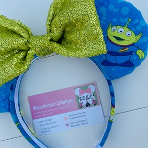Pu&ograve; includere: Una fascia per capelli a tema Toy Story con orecchie blu stampate con un personaggio alieno e un grande fiocco di paillettes verde lime. La fascia ha un interno bianco e un biglietto da visita con il nome del marchio.