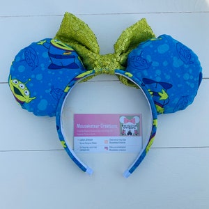 Pu&ograve; includere: Orecchie da topo con un tessuto blu con una stampa aliena e un fiocco di paillettes verde lime. La fascia &egrave; bianca con una stampa aliena abbinata. L'immagine include un biglietto da visita con il testo "Mouseketear Creations".