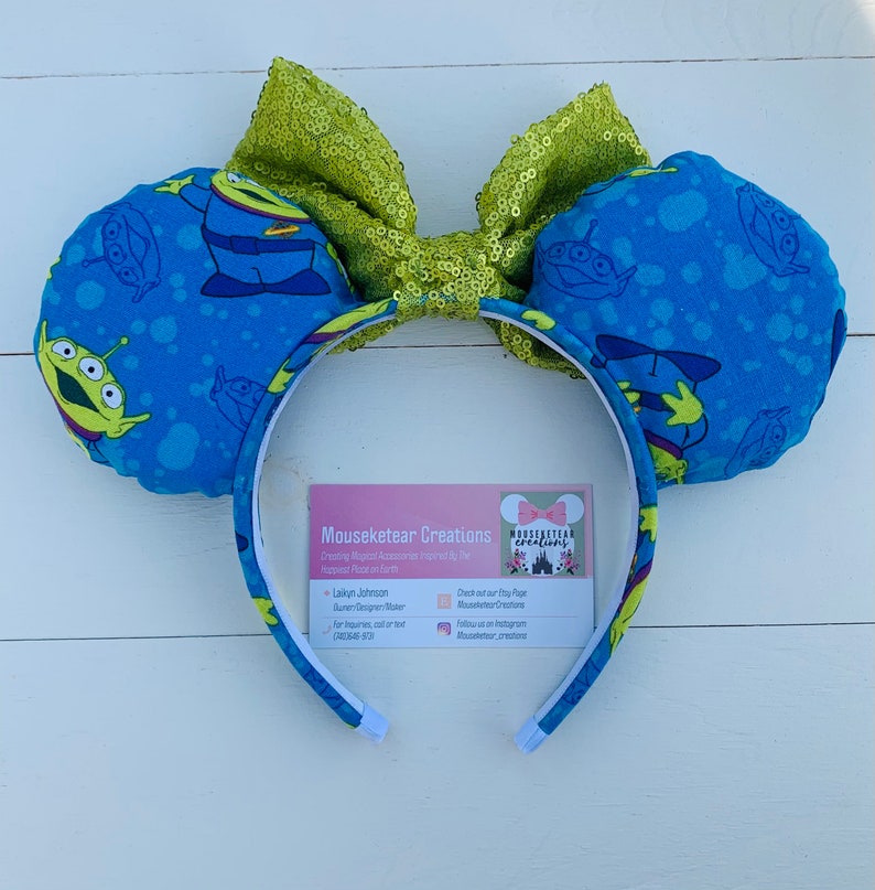 Pu&ograve; includere: Orecchie da topo con un tessuto blu con una stampa aliena e un fiocco di paillettes verde lime. La fascia &egrave; bianca con una stampa aliena abbinata. L'immagine include un biglietto da visita con il testo "Mouseketear Creations".