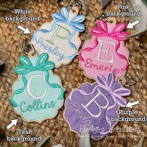 May include: Embroidered keychains in white, pink, teal, and purple. Each keychain features a letter and name, with a bow and ribbon design. The keychains are attached to silver-colored key rings. The text "Kimbo's Kreations" is at the bottom.