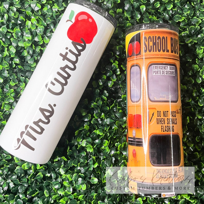 May include: Two stainless steel tumblers. One is white with black text that says "Mrs. Curtis" and a red apple graphic. The other is yellow with a school bus graphic and the text "SCHOOL BUS" and "EMERGENCY DOOR PORTE DE SECOURS" and "DO NOT PASS WHEN SIGNALS FLASHING".