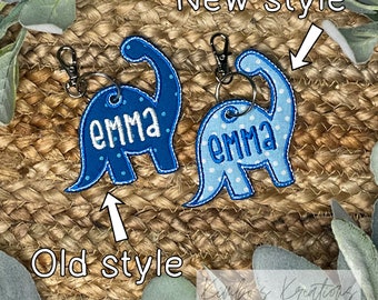 May include: Two blue dinosaur-shaped keychains with the name "Emma" embroidered on them. The keychain on the left is the "old style" and the keychain on the right is the "new style".